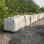 Long row of bulk fertilizer bags on pallets along the wood line at a farm for agricultural use.